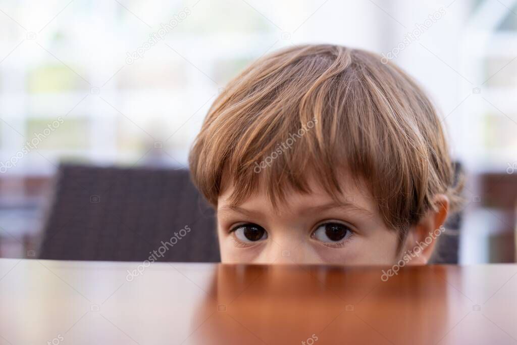 Fearful little boy hide half face under wooden table in cafe hall closeup, free copy space. Pretty kid of kindergarten age scared to sit in kitchen. Morning, breakfast, phobia, alone.