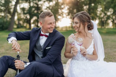 Yazın parkta çimlerin üzerinde oturan neşeli düğün çifti portresi. Genç adam şarap şişesini açıyor, genç ve güzel bir kadın gelin gözlük tutuyor, gözleri buluşuyor. Kutlama, ilişki.