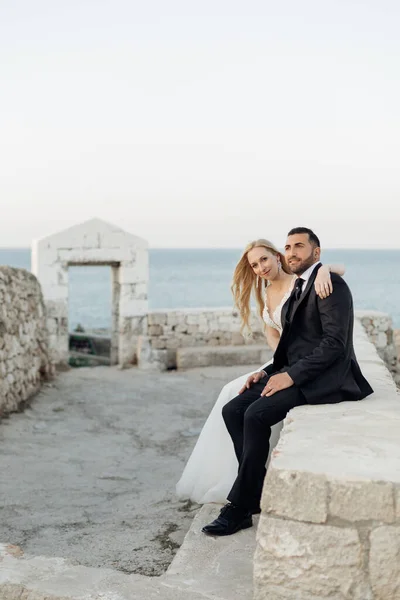 Portrait of happy wedding couple sitting on coast of Adriatic sea on ancient stones. Young woman bride with fair hair wearing long white wedding dress, putting arm around grooms neck in suit. Wedding.