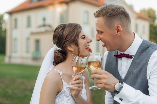 Side view of funny wedding couple standing outside near old building in park in summer, showing tongues to each other. Young man groom and woman bride raising hands with glasses of wine. Celebration.