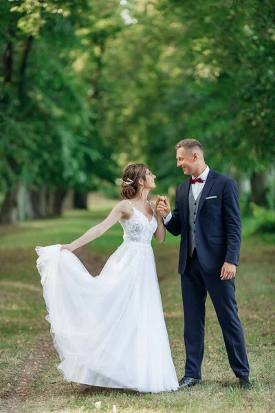 Side view of loving wedding couple standing outside in park in summer. Young man groom holding hand of young amazing woman bride, dancing, looking at each other. Relationship, love, celebration.