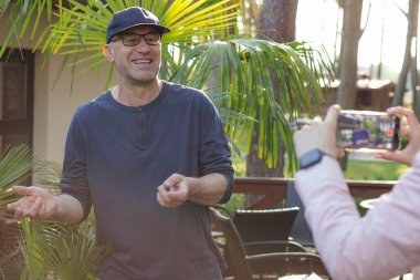 Şapkalı, gözlüklü, neşeli bir adam canlı yayında gülüyor ve el kol hareketi yapıyor. Kırpılmış kadın eller, yurtdışındaki gezginlerin rekor seviyede eğitim programı için akıllı telefonu tutuyor. Vlog, yayınlanan hikayeler çevrimiçi