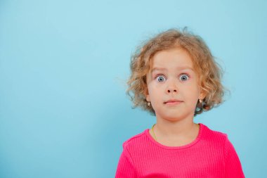 Küçük anlamlı, şok olmuş, korkmuş, pembe tişörtlü kıvırcık sarışın kız mavi izole stüdyo arka planında kameraya bakıyor. Aile sorunu, taciz, zorbalık. Reklam ve afiş. Boşluğu kopyala