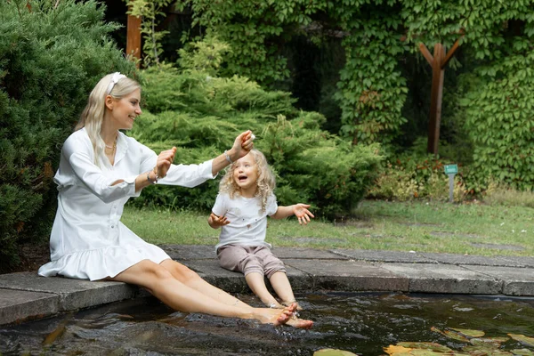 Smiling barefoot blonde woman and little curly girl raising arms and washing legs in lake water in natural park in hot summer. Family holidays and vacation. Bound with nature. Children care and safety