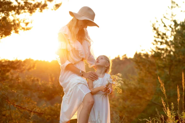 Adorable, pretty, charming, joyful, calm woman, mother in hat and little blonde girl, daughter in dress with flower herbs, have fun on meadow in sunny natural forest area. Mother day celebration
