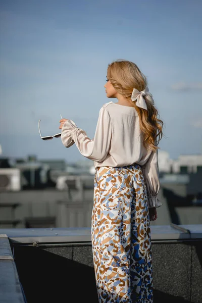 Glamorous young woman in elegant blouse and patterned pants stand on roof terrace with sunglasses back view. Photo of parisian lady in stylish outfit on sky background. Paris fashion, roof, sky.