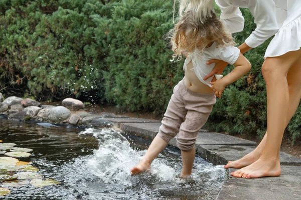 Unrecognizable woman holding blonde little girl, helping to wash legs in lake water in natural park in hot summer in the countryside. Relaxation and bound with nature. Parenthood help, protection