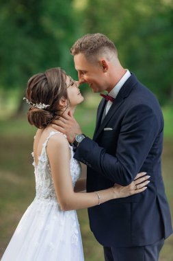 Mutlu evlilik çifti yazın parkta dikilirken yan görüntüsü. Genç damat kollarını genç, güzel bir kadının boynuna doladı, kucakladı, gözleri gördü, öptü. İlişki, aşk.