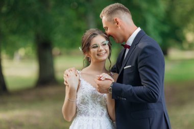 Yazın parkta dikilen gülümseyen düğün çifti portresi. Genç ve güzel bir kadın gelin genç bir damadın elini tutuyor, sarılıyor, sarılıyor. İlişki, aşk, ilgi, kutlama.