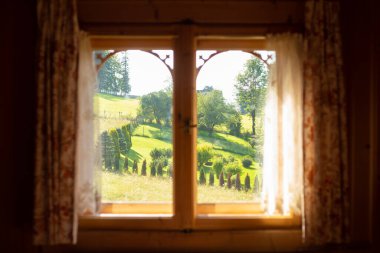 Perdeleri olan ahşap pencerenin önü, güneşli kırsal manzarası ağaçlar ve tepeler. Kırsal tatiller, temiz hava ve farkındalık. Seyahat ve tatil doğada. Kapalı bir ev.