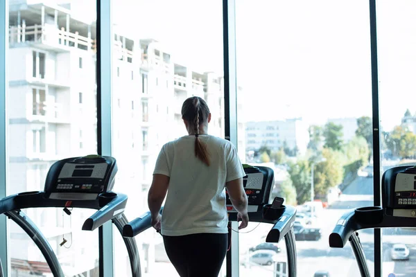 Fat woman alone run on treadmill at fitness gym and look out window at transport traffic and city, back view. Overweight lady do cardio workout on training apparatus. Weight loss, sport lifestyle.