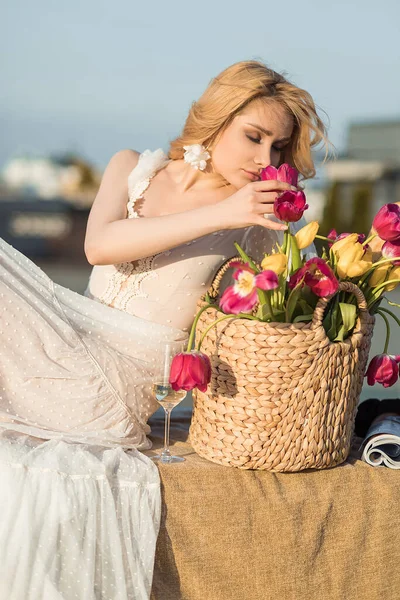 Charming woman in romantic dress smell bouquet of pink and yellow tulips in wicker basket on roof terrace. Elegant lady relax with glass of champagne. Paris style, roof, sky, stylish decor.