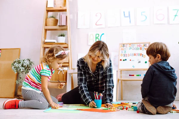 Kind teacher conduct lesson for little children while sitting on floor in playing room. Kids play with toys and draw picture with colored pencils in kindergarten. Lesson, preschool, daycare.