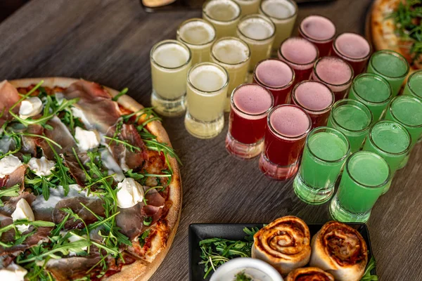 Pizza with mozzarella cheese, bacon, meat on cafe table. Alcohol colorful red, yellow and green jelly drinks and cinnamon rolls or buns as dessert. Puff pastry rolls with filling and meat with sauce