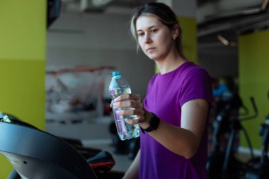 Spor salonunda koşu bandında koşan terli bir kız ve bir şişe su. Ayrıca beden ölçüsünde bir kadın egzersiz makinesinde egzersiz yapıyor. Kilo kaybı, susuzluk, spor salonu üyeliği, spor yaşam tarzı..