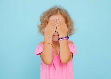 Kısa kıvırcık saçlı, pembe tulumlu, bilezik takan, mavi arka planda yüzünü saklayan elleri olan üzgün küçük bir kızın portresi. Duygular, korkular, saklambaç, stüdyo.