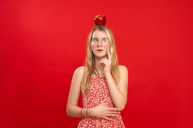 Şaşkın, güzel, sarışın bir kızın portresi. Gözlük takan büyük kırmızı bir elma. Kırmızı arka planda işaret parmağıyla yanağa dokunuyor. Stüdyo. Fikir, aydınlatma, deha, kopyalama alanı.