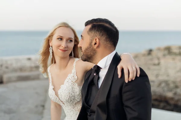 Portrait of loving wedding couple sitting on coast of Adriatic sea, meeting eyes. Young woman bride with fair hair wearing long white wedding dress, putting arm around grooms neck in suit. Wedding.