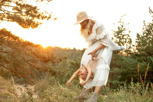 Portrait of laughing family wearing white dresses, walking in park forest illumined by sunset in summer. Young gorgeous woman mother holding upside down little girl daughter, playing. Relationship.