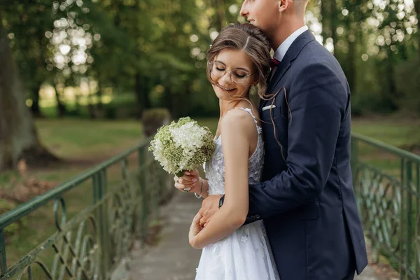 夏に公園の橋の上に立って陽気な結婚式のカップルの肖像画 閉じた目で花嫁介添人花束を保持若い完璧な女性 後ろから花嫁を受け入れる若い新郎 関係愛 — ストック写真