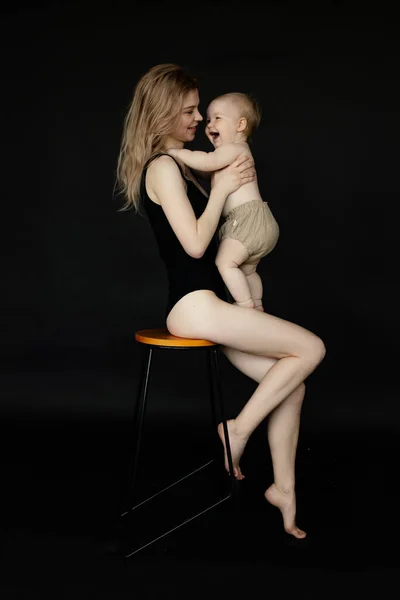 Vertical portrait of young blonde barefoot woman, mother in black bodysuit embracing and playing with toddler baby on black background. Children care and breastfeeding. Maternity vacation