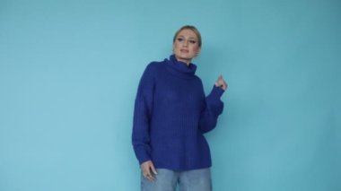 Sakin, rahat, neşeli, gülümseyen, mavi, sıcak, büyük, modern süveterli sarışın kadın mavi stüdyo arka planında dans ediyor. Rahat ve rahat doğal giysiler. Sonbahar ve kışa hazırım. Uzayı kopyala 4k
