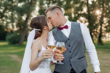 Yazın parkın dışında bekleyen harika bir çiftin yan görüntüsü. Genç damat, genç ve çekici gelini öpüyor, şarap kadehleriyle ellerini uzatıyor. İlişki, aşk, kutlama..