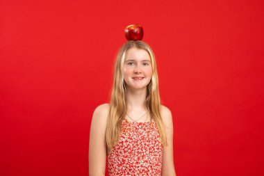 Büyük kırmızı elmayı kırmızı arka planda dengede tutan uzun sarı saçlı, gülümseyen genç kızın portresi. Stüdyo. Fikir, aydınlanma, dersler, eğitim, deha, sağlık, kopyalama alanı.