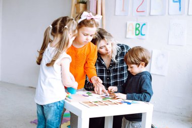 Sarışın bir kadın, öğretmen ve küçük çocuklar oynayıp öğreniyorlar. Anaokulu çocukları için çocuk yetiştirme merkezi kuruyorlar. Anaokulu ve gündüz erken eğitim. ABC, mantık, matematik sınıfı