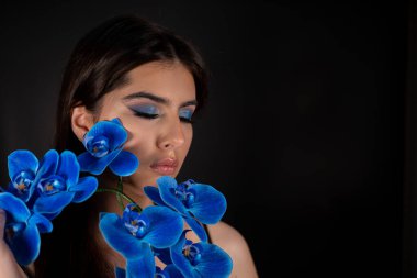 Çekici, sakin, rahat, siyah saçlı, makyajlı, mükemmel tenli, mavi gözlü, boynunda ve omuzlarında mavi süs çiçekleri olan bir kadın. Kozmetik ve makyaj dersi. Boşluğu kopyala