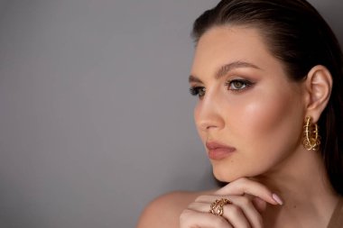 Modayı takip eden, modaya uygun, eli çenesinde, profesyonel makyajlı, gri arka planda kameradan uzağa bakan şık esmer kadın yüzü. Lüks klasik tasarım süsü. Boşluğu kopyala