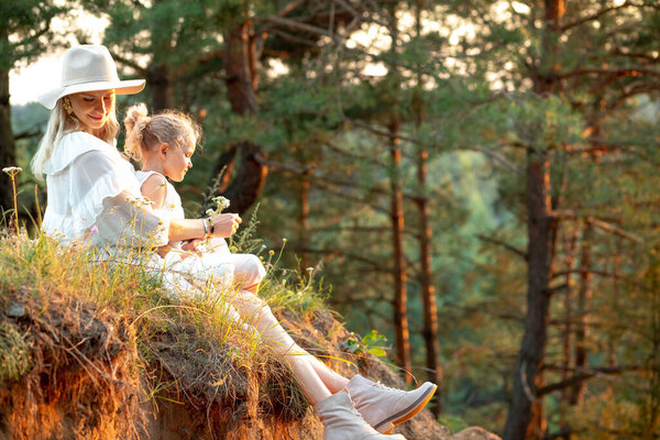 Adorable woman, mother in hat and little blonde girl in dress sitting together on slope in sunny natural forest area. Ecological vacation and holiday with family, healthy active lifestyle. Copy space