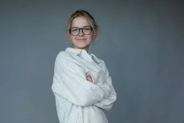 Smiling young woman in black glasses get warm by crossing arms, gray background, free copy space. Cozy white sweater, shirt for cold weather. Casual seasonal clothes, female basic wear.