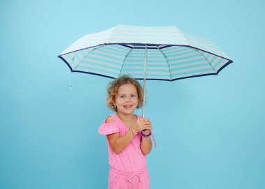 Mavi gözlü, kıvırcık saçlı, pembe tulumlu, mavi arka planda açık çizgili şemsiye tutan neşeli bir kız portresi. Hayat sigortası, güvenlik, sigorta, kopyalama alanı, koruma.