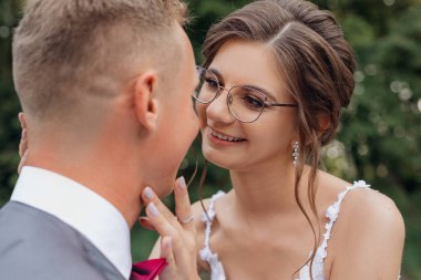 Ağacın yanında duran neşeli düğün çifti portresi. Genç, harika, gülümseyen bir kadın gelin parmaklarıyla damadın boynuna dokunuyor, gözleri buluşuyor, poz veriyor. İlişki, aşk, düğün..