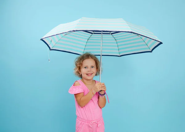 明るい青の肖像画 青い背景にオープンストライプの傘を保持し ピンクのジャンプスーツを着て巻き公正な髪を持つ少女目 生命保証 安全性 コピースペース — ストック写真
