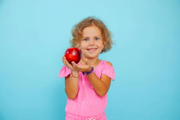 Portrait of amazing blue-eyed little girl with curly fair hair wearing pink jumpsuit, holding showing big red ripe apple on blue background. Studio. Healthy food, fruits, vitamins, summer, ecology.