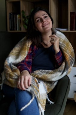 Dikey, olgun, sakin, dişlek gülüşlü, battaniyeyle örtülü, kameraya bakan ve kapalı bir kütüphanedeki koltukta oturan esmer kadın. Hafta sonu aktivitesi ve sıcak kıyafetlerle rahat bir gün.