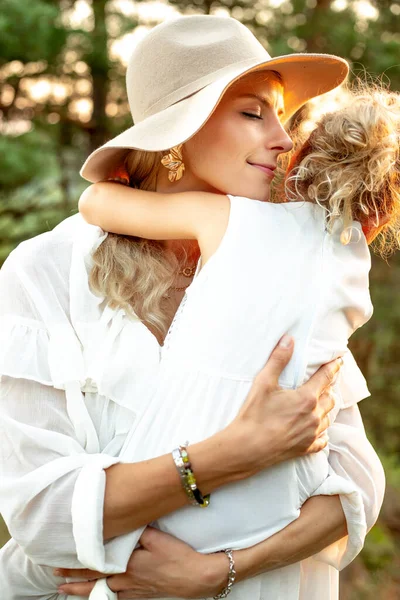 Vertical charming, kind, loving stylish woman in hat hugging little blonde girl, daughter with close eyes on walk trip in sunny wood forest. Holiday with family, healthy lifestyle. Adoption and care