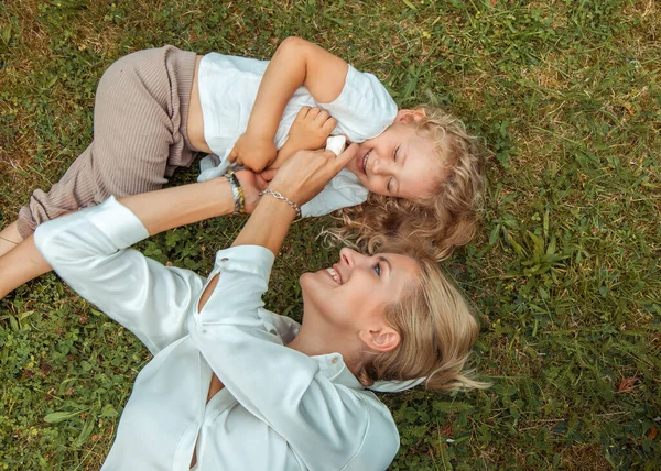 Close up shiny, laughing, kind, soft blonde family of mother playing, tickling little blonde daughter, spending time together and lying on fresh green grass meadow in sunny natural area. Top view