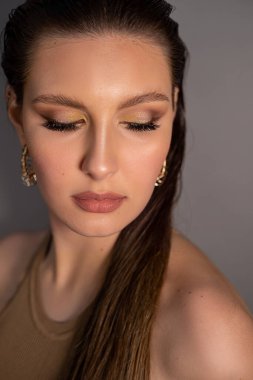 Dikey moda, zarif, parlak, gösterişli esmer kadın yüzü, profesyonel makyaj, altın aksesuarlar, gri arka planda aşağı bakmak. Bayramlarda kadınlar için görkemli bir hediye. Düğün hediyeleri.