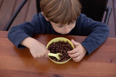 Küçük çocuk mutfakta, kahverengi ahşap masada oturuyor ve kahvaltı manzarası seyrediyor. Anaokulu çocuğu elinde kaşık tutarak çikolatalı topları sütle yiyor. Kahvaltı hazır, sağlıklı beslenme.