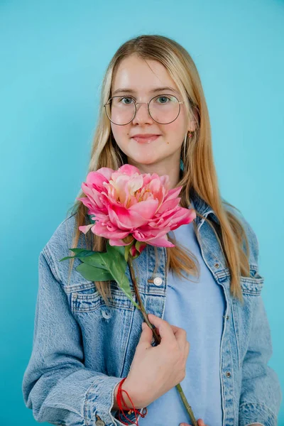 可爱的少女抱着芬芳的粉红色鲜花 长长的茎 蓝空的背景 戴眼镜快乐的年轻女士 笑着欣赏盛开的植物的美丽 艳丽的花朵概念 — 图库照片