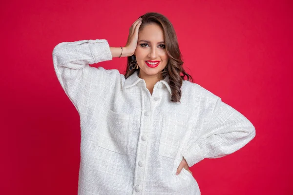 Confident, pretty, smiling plump woman with red lipstick toothy smile to camera in white oversize shirt, hold hand in hair on red studio background. Xl size, plus size and self acceptance. Copy space