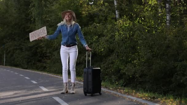妇女站在森林里等待路过的汽车 路边挂着行李箱和纸板海报 戴帽子的金发女士从快乐中跳到任何地方 搭便车 — 图库视频影像