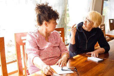 İlgili iki yaşlı kadının portresi gündelik giysiler giymiş, ahşap masada oturmuş, tartışıyorlar, not defteriyle notlar alıyorlar, güneşli büyük oda ofisinde el kol hareketi yapıyorlar. Eğitim, ağ pazarlaması, planlar.