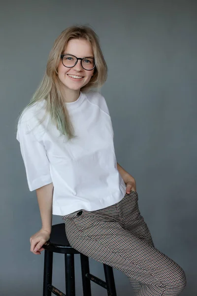年轻迷人的年轻女子的画像 长长的金发 身穿白色T恤 灰色裤子 坐在黑色凳子上 靠在手上 在灰色背景上摆姿势 工作室 — 图库照片
