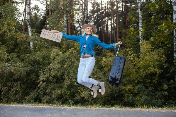 Portrait of young happy woman wearing blue shirt, white jeans, beige floppy hat, holding cardboard with inscription anywhere and handle of black suitcase, jumping. Travelling, hitchhiking, levitation.