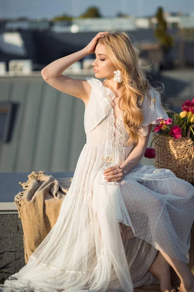 迷人的女人穿着时髦的白色衣服坐在毛毯上 回头望着屋顶阳台上的落日 优雅的女士喝杯香槟放松一下 巴黎时尚 — 图库照片