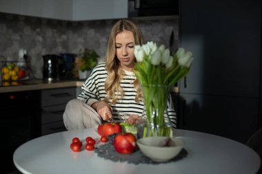 Ev kıyafetleri içinde sevimli bir sarışın mutfaktaki masasında oturuyor, önünde taze sebzeler ve meyveler var ve beyaz laleli vazo var. Sağlıklı bir yaşam tarzı. Organik yiyecekler. 